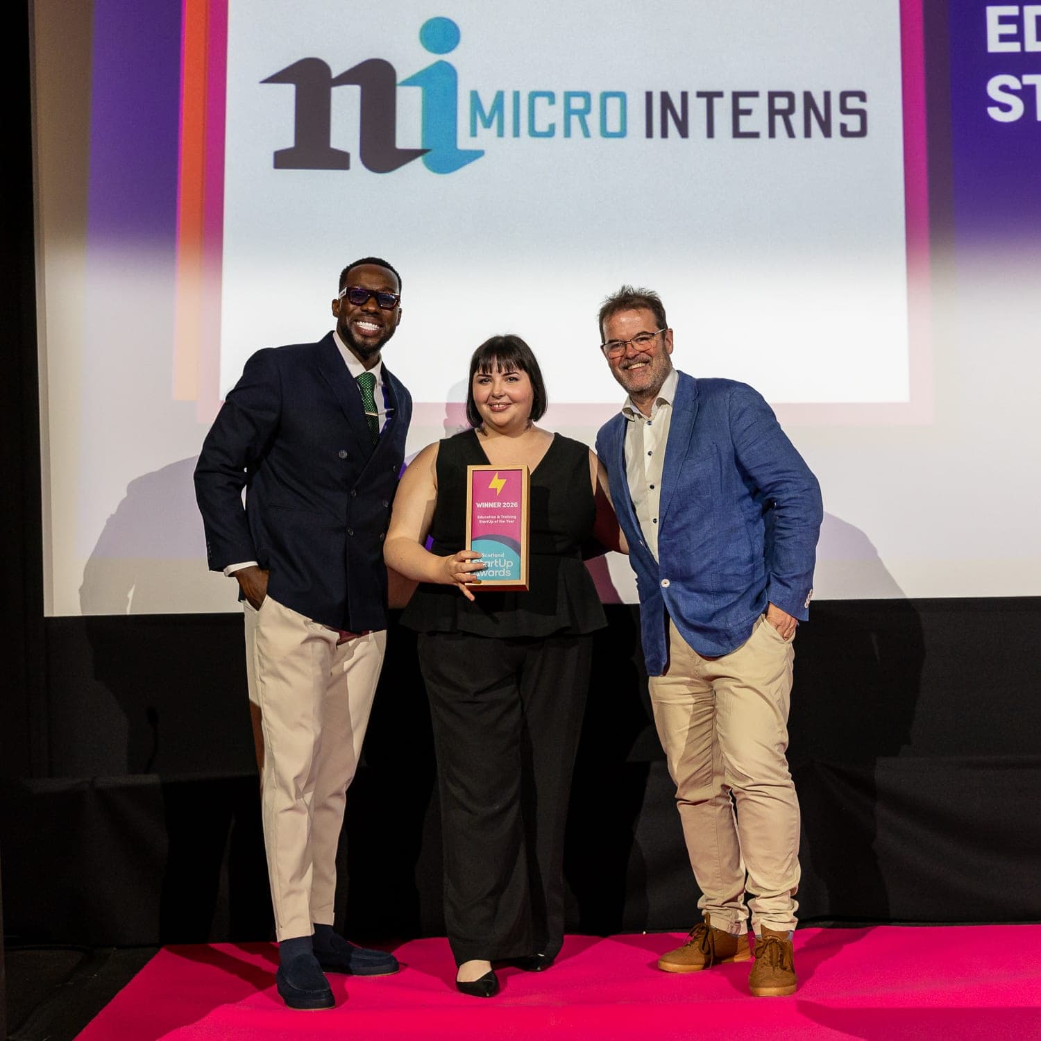 The MicroInterns founder on stage at the UK StartUp Awards Scotland final, holding the Winner 2026 Education & Training StartUp of the Year trophy between the two award presenters, the MicroInterns logo on the screen behind them