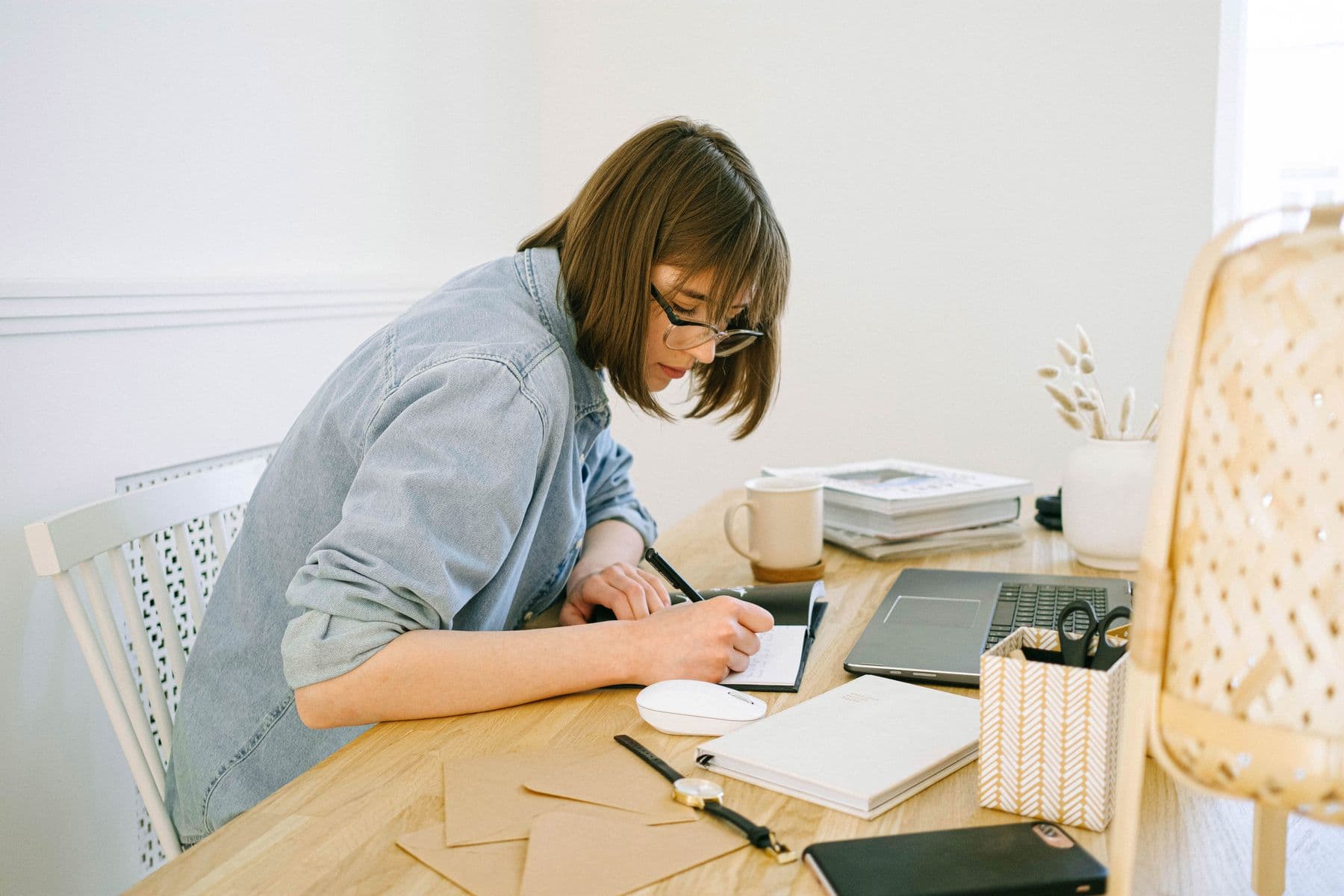A student bent over a notebook at a light wooden table, drafting an application beside a laptop, a coffee and a fan of unsealed envelopes