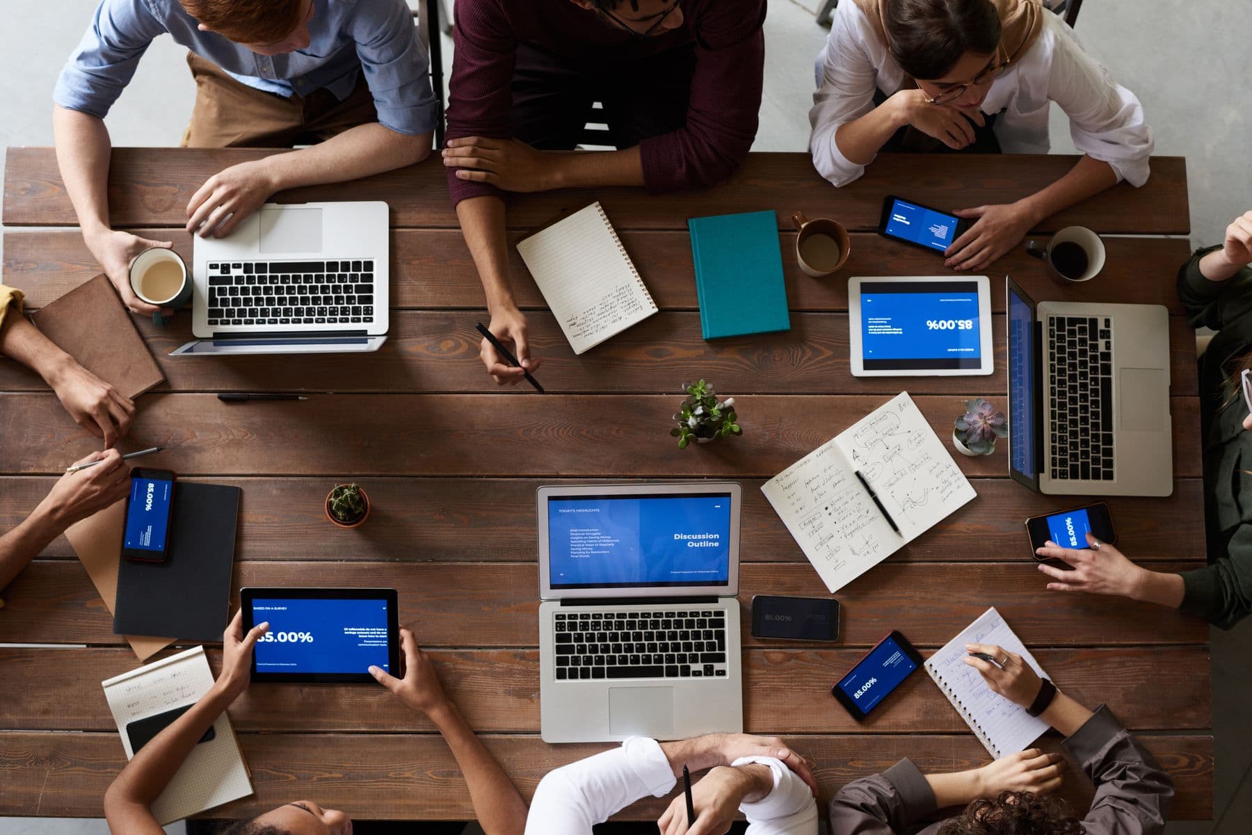 Looking down on a long wooden table where a team is working together, laptops, notebooks and coffee spread between them