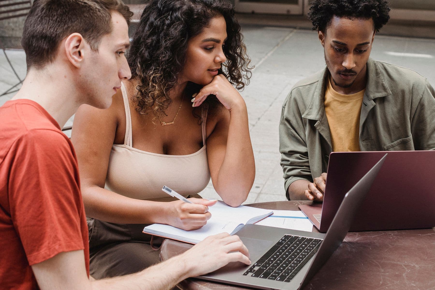 Three students comparing notes around laptops at an outdoor table
