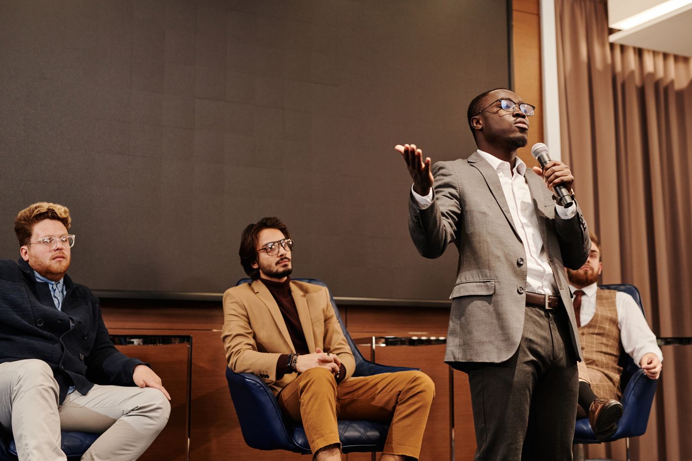 A speaker on a panel gesturing mid-answer, three other panellists seated beside him in front of a seated audience